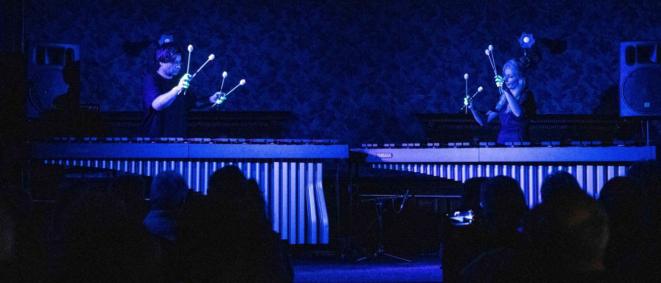 Zwei Personen spielen auf einer Bühne mit Musikinstrumenten unter blauem Licht vor einem Publikum.