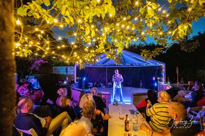Eine nächtliche Outdoor-Veranstaltung unter einem Zelt mit Lichterketten, ein Mann auf der Bühne singt mit einem Mikrofon und ein Publikum sitzt um einen Tisch mit Getränken.