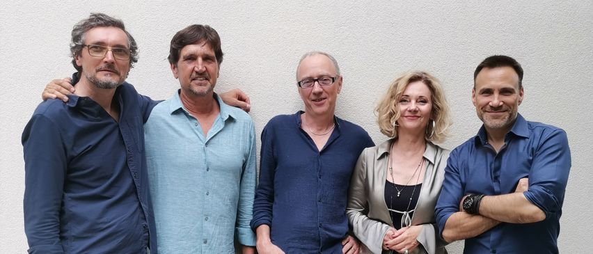 Fünf Personen stehen eng beieinander vor einer weißen Wand. Sie lächeln und posieren für ein Foto. Jede Person trägt Freizeitkleidung.