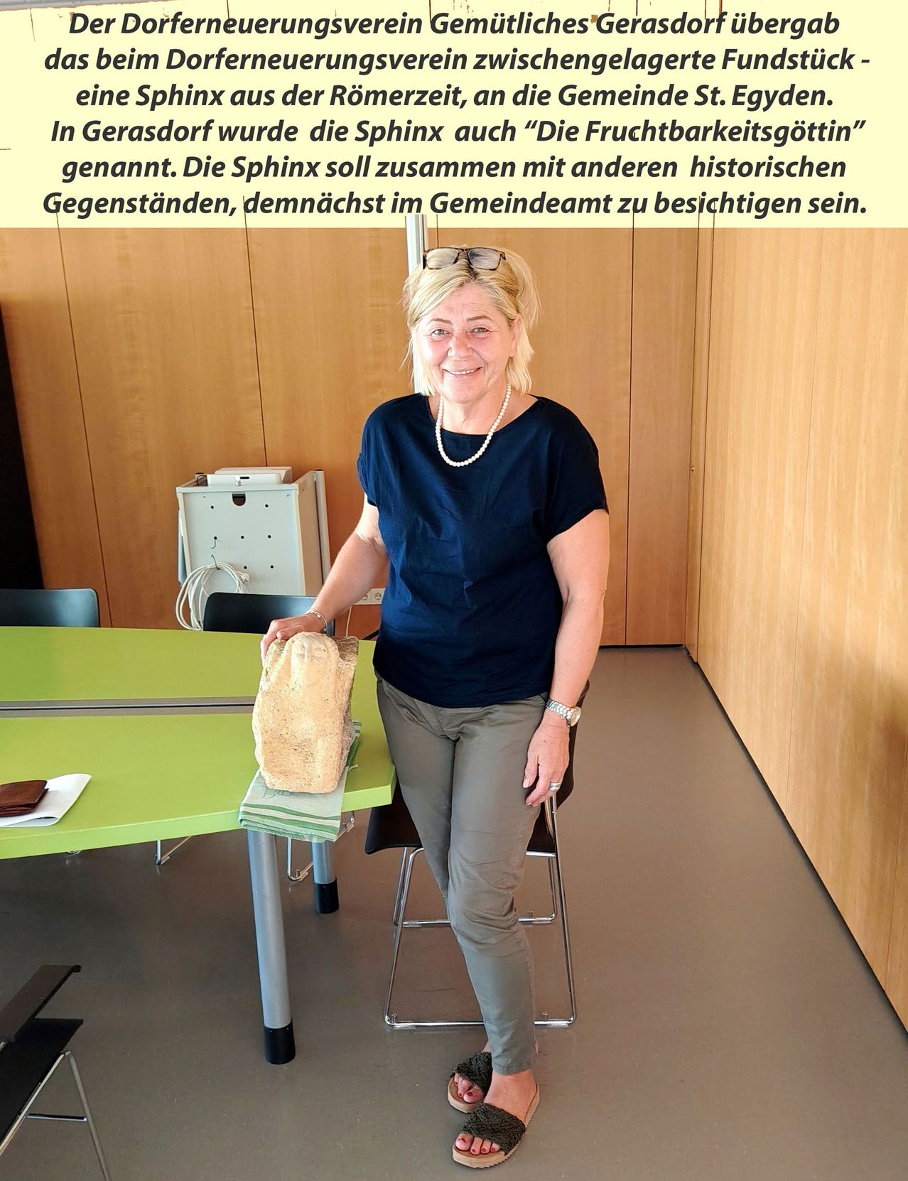 Eine Frau in einem blauen Shirt und grauen Hosen steht in einem Büroraum und hält eine Steinskulptur einer Sphinx. Der Raum hat Holzwände und einen grünen Schreibtisch.
