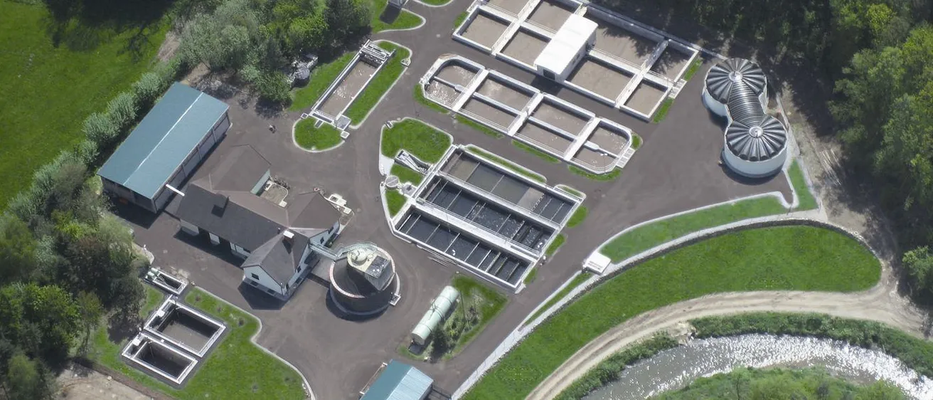 Ein Luftbild einer Kläranlage mit mehreren großen Tanks und rechteckigen Teichen. Ein Gebäude befindet sich links und ein Fluss rechts. Grüner Rasen und Bäume umgeben das Gebiet.