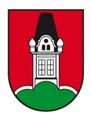Das Bild zeigt ein Wappen mit einem schwarzen dreieckigen Turm auf einem roten Schild. Der Turm hat ein spitzes Dach und steht auf einem grünen Hügel. Das Wappen ist von einem schwarzen Rand umrahmt.