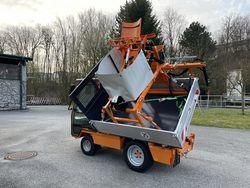 Ein orangefarbener Maschinenwagen mit angehobener Rückenlehne steht auf einem gepflasterten Bereich, mit einem Steingebäude und Bäumen im Hintergrund.