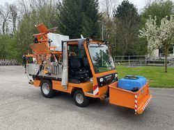 Ein orangefarbener Nutzfahrzeug mit einer Sprühvorrichtung und einem großen blauen Tank an der Seite ist auf einer gepflasterten Oberfläche geparkt. Im Hintergrund befinden sich Bäume und eine Rasenfläche.