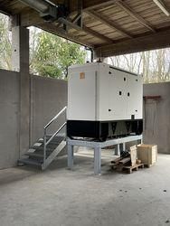 Ein großer weißer und schwarzer Industriegenerator steht auf einem Betonboden. Er befindet sich auf einem Metallständer. Links sind Holztreppen und rechts einige Holzpaletten und ein Holzkasten. Der Raum besteht aus Betonwänden und hat eine Holzdecke. Draußen sind Bäume zu sehen.