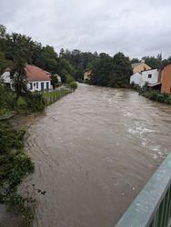 Ein überfluteter Fluss fließt durch eine kleine Stadt mit Wohnhäusern und Bäumen am Ufer.