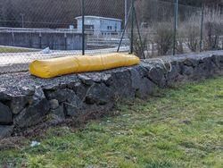 Eine gelbe Schutzabdeckung liegt auf einer Steinmauer, gesichert mit einem Seil. Hinter der Mauer umschließt ein Zaun einen Grasbereich mit einem Gebäude in der Ferne.