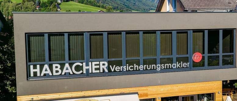 Ein Gebäude mit einem Schild, das 'HABACHER Versicherungsmakler' steht. Es hat große Fenster, einen Balkon und ein kleines Schild mit dem Wort 'Café' auf der rechten Seite. Das Gebäude ist von grünen Hügeln und Bergen umgeben.