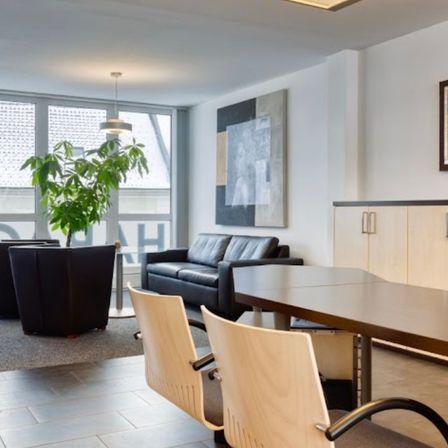 Ein modernes Bürozimmer mit einem schwarzen Sofa, zwei Sesseln und einem langen Holztisch. Eine Pflanze, eine hängende Lampe und ein gerahmtes Bild befinden sich an der Wand. Der Raum hat ein Glasfenster mit Jalousien und einen gefliesten Boden.