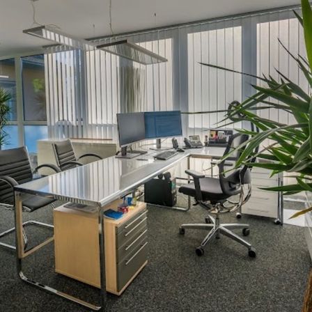 Ein Büroraum mit einem großen Schreibtisch, zwei Monitoren und einem Telefon. Zwei schwarze Bürostühle befinden sich auf beiden Seiten des Schreibtischs. Eine Pflanze steht rechts.