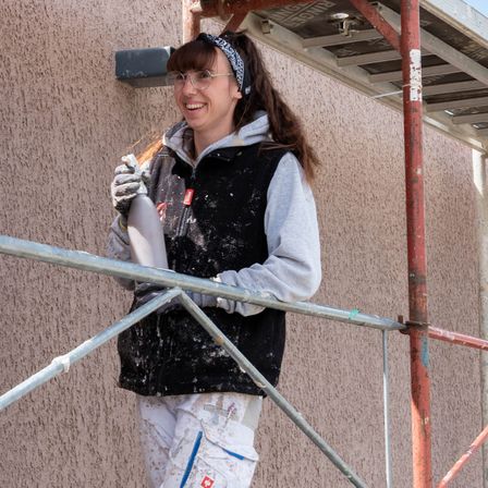 Eine Frau in einem schwarzen Weste und Brille steht auf einem Gerüst, lächelt und hält ein silbernes Objekt in der linken Hand. Sie scheint etwas an der Wand zu arbeiten.