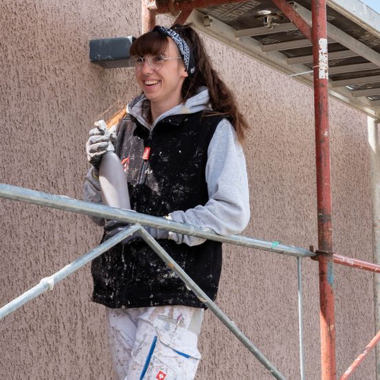 Eine Frau in einem schwarzen Weste und Brille steht auf einem Gerüst, lächelt und hält ein silbernes Objekt in der linken Hand. Sie scheint etwas an der Wand zu arbeiten.