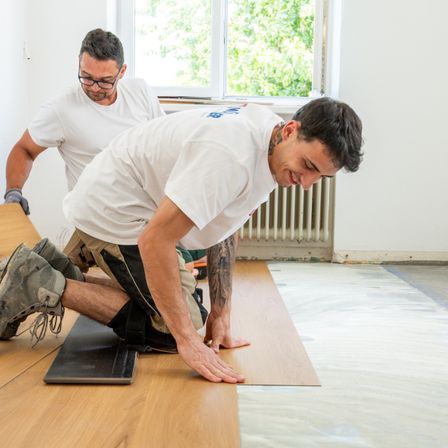Zwei Männer in weißen T-Shirts und Handschuhen arbeiten an Holzböden. Einer kniet, lächelt, während der andere in der Nähe steht. Sie sind in einem Raum mit Fenster und Heizkörper.