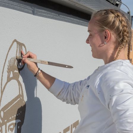 Eine Frau mit geflochtenen Haaren malt eine Wand mit einem Pinsel. Sie trägt ein weißes T-Shirt und hat Armbänder am Handgelenk. Die Wand hat einen Schatten.