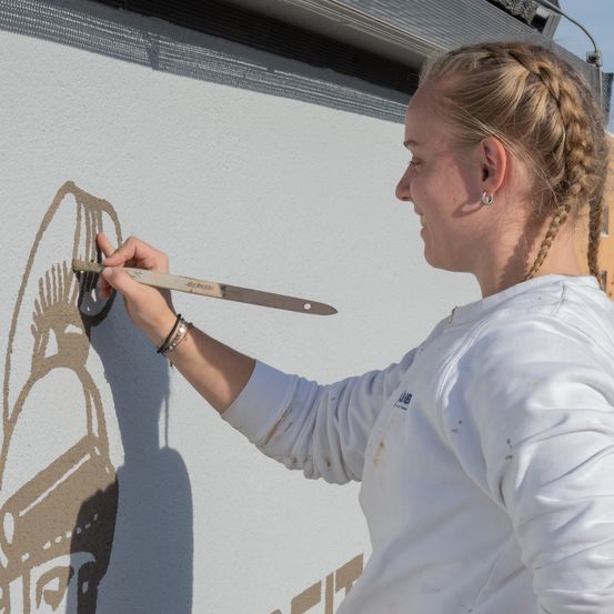 Eine Frau mit geflochtenen Haaren malt eine Wand mit einem Pinsel. Sie trägt ein weißes T-Shirt und hat Armbänder am Handgelenk. Die Wand hat einen Schatten.