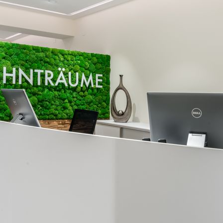 Ein moderner Büroempfangsbereich mit zwei Dell-Monitoren, einem Stuhl und einem Schreibtisch mit einer Vase. Eine grüne Mooswand zeigt das Wort 'HinTräume'.