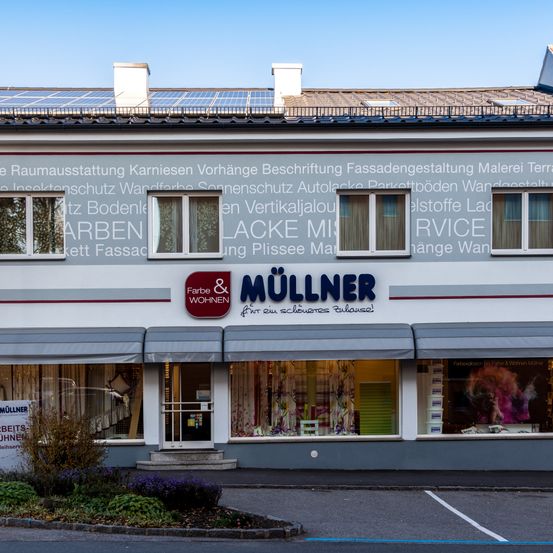 Ein Ladengeschäft mit grauen Vordächern und Fenstern zeigt das Schild 'MULLNER' und 'Farbe & Wohnen'. Solarmodule sind auf dem Dach. Das Gebäude hat mehrere Fenster mit Vorhängen.