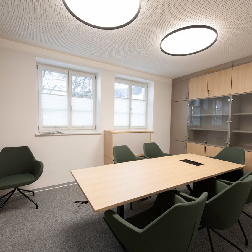 Ein leerer Besprechungsraum mit einem langen Holztisch, grünen Stühlen, zwei großen Fenstern und zwei runden Deckenleuchten.