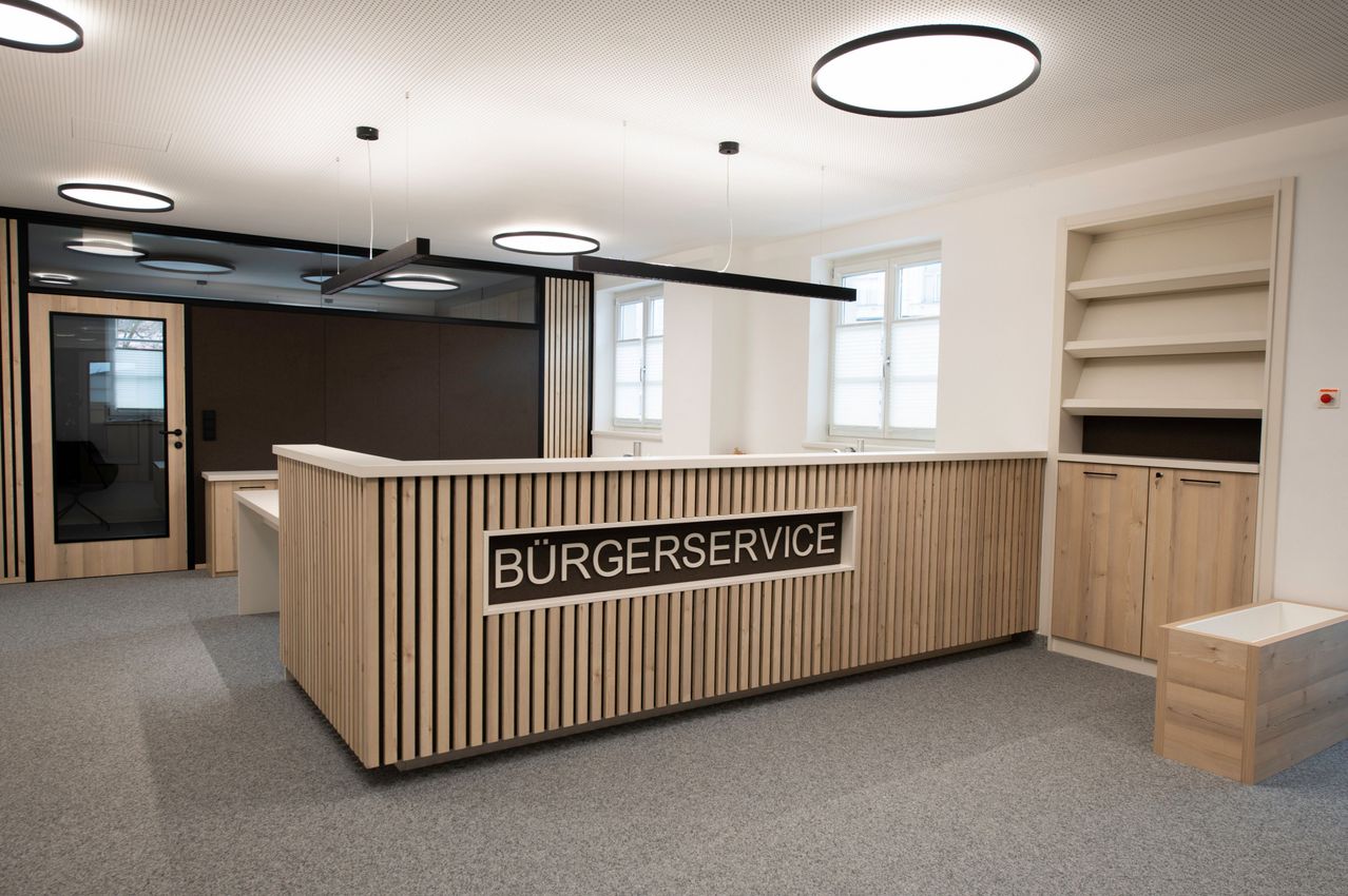 Eine Empfangstheke in einem modernen Büroraum mit Holzlamellen und einem Schild, das Burgerservice steht. Die Decke hat kreisförmige Beleuchtung.