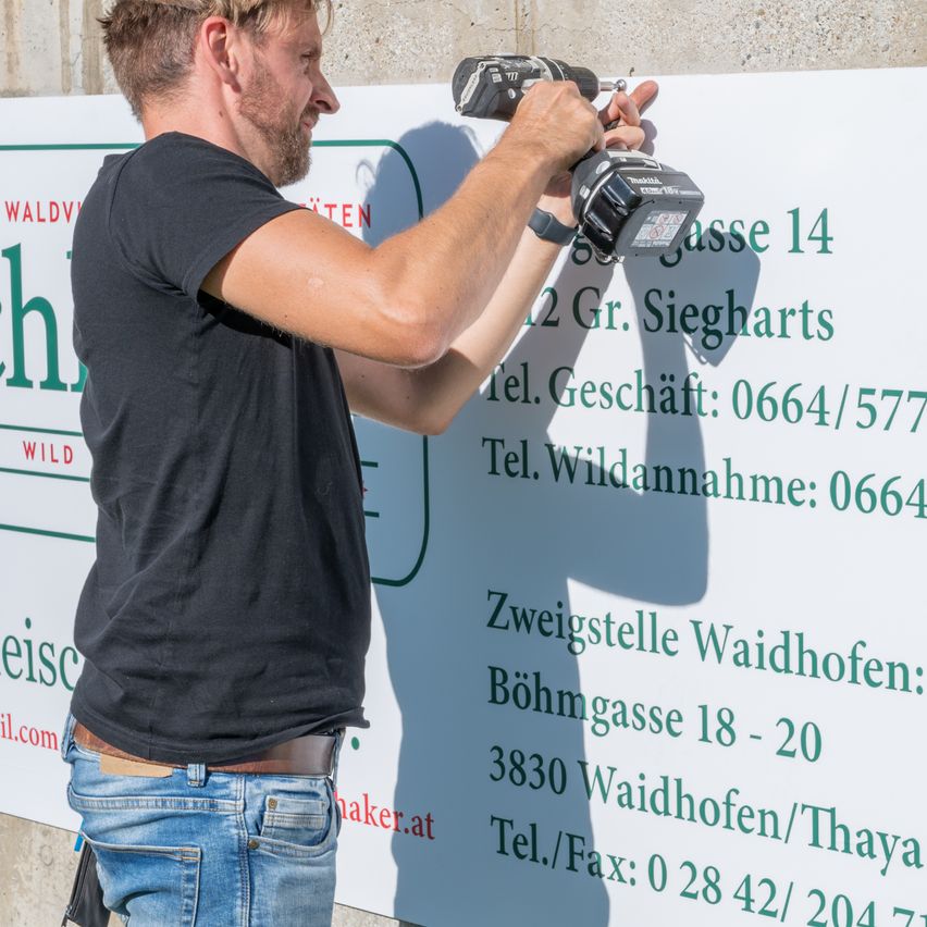 A man wearing a black shirt and jeans is drilling a wall. Behind him is a large sign with phone numbers and addresses.