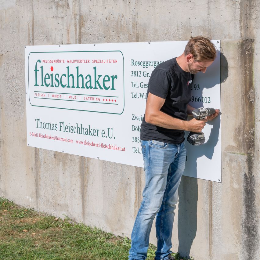 A man is fixing a sign with the text 'fleischhaker' on a concrete wall. The sign includes details like address, phone numbers, and website.