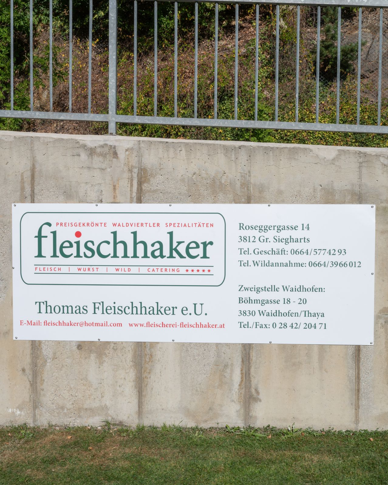 A white sign mounted on a concrete wall with the text 'Fleischhaker' and other details about the business. The address, phone numbers, and website are included.