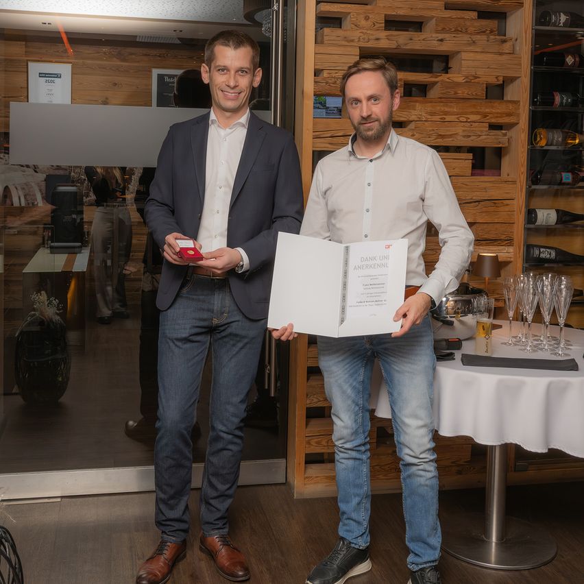 Zwei Männer stehen drinnen in der Nähe einer Glastür, einer hält ein Zertifikat und eine rote Box, der andere hält ein großes offenes Zertifikat. Hinter ihnen eine holzverkleidete Wand mit Weinflaschenregalen. Im Hintergrund ein Tisch mit Weingläsern und einer Lampe.