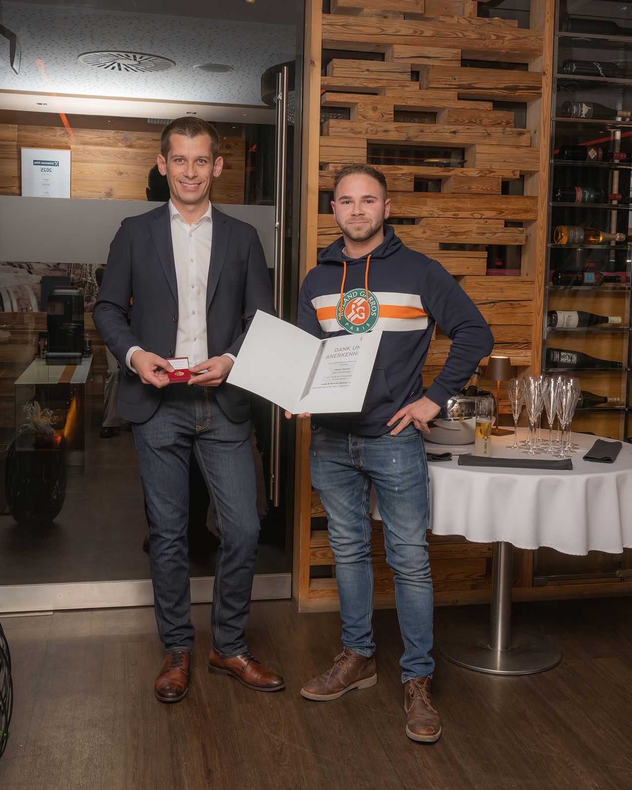 Zwei Männer stehen in einem Raum und halten ein Zertifikat und eine kleine Schachtel, lächelnd. Dahinter steht ein Tisch mit Weingläsern, einer Lampe und Flaschen. Eine Tafel und ein Regal mit Gegenständen sind an der Wand.