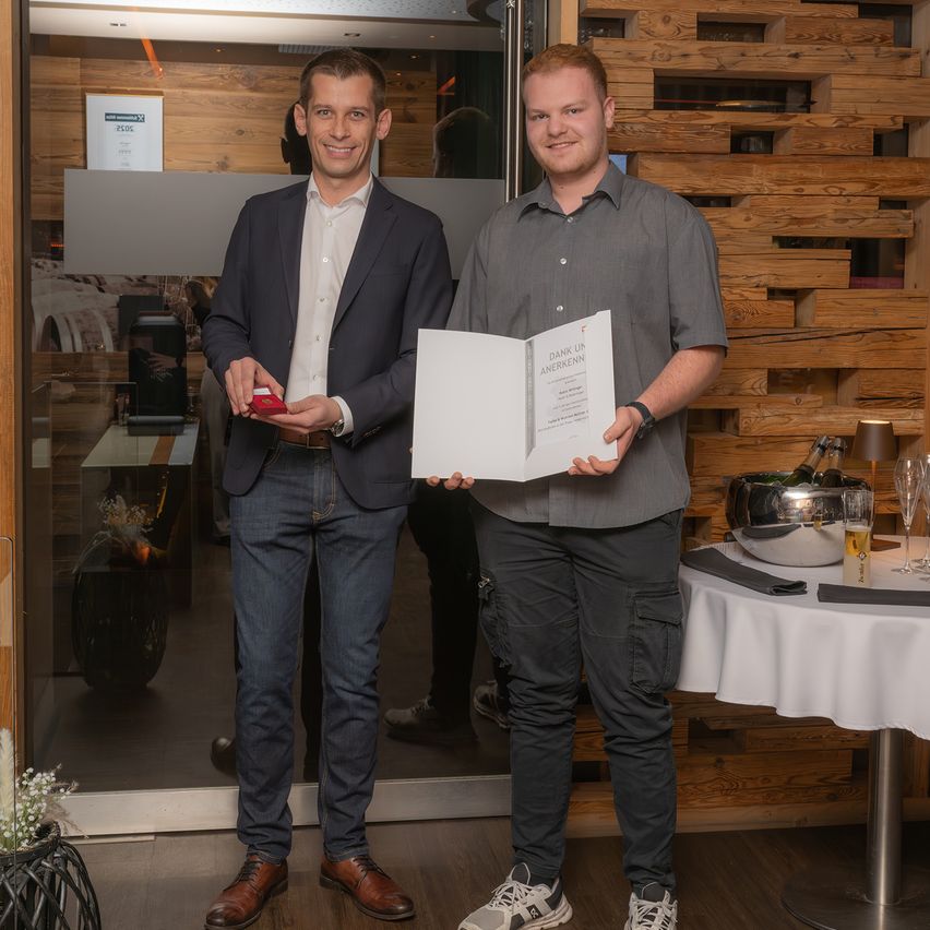 Zwei Männer stehen in einem Raum, einer hält ein Buch und der andere eine kleine rote Box. Hinter ihnen befindet sich eine Glastür mit einem Schild und einem Tisch mit einer Schüssel, einer Lampe und Weingläsern.