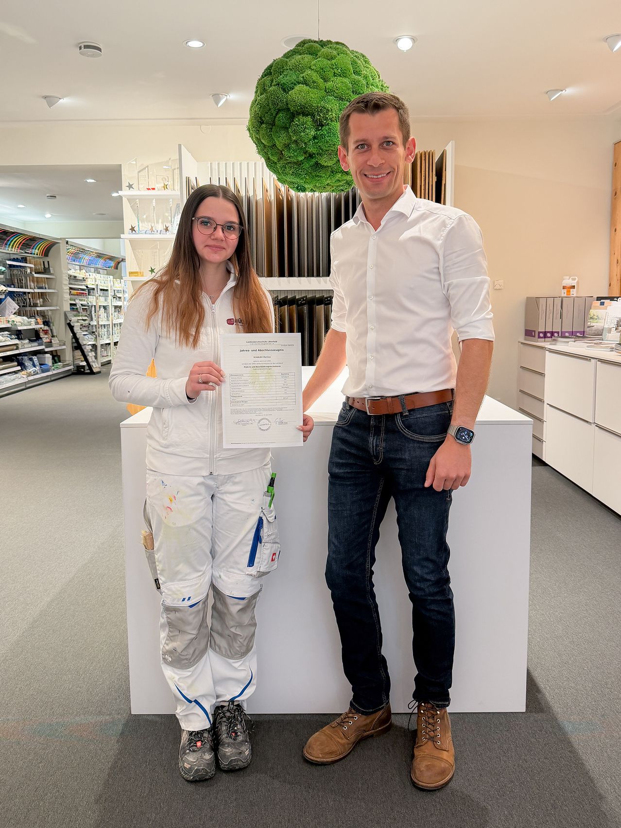 Eine Frau in einer besprühten Kleidung hält ein Zertifikat, während sie neben einem lächelnden Mann in einem weißen Hemd und Jeans steht. Sie sind in einem Raum mit Regalen und einer weißen Theke.