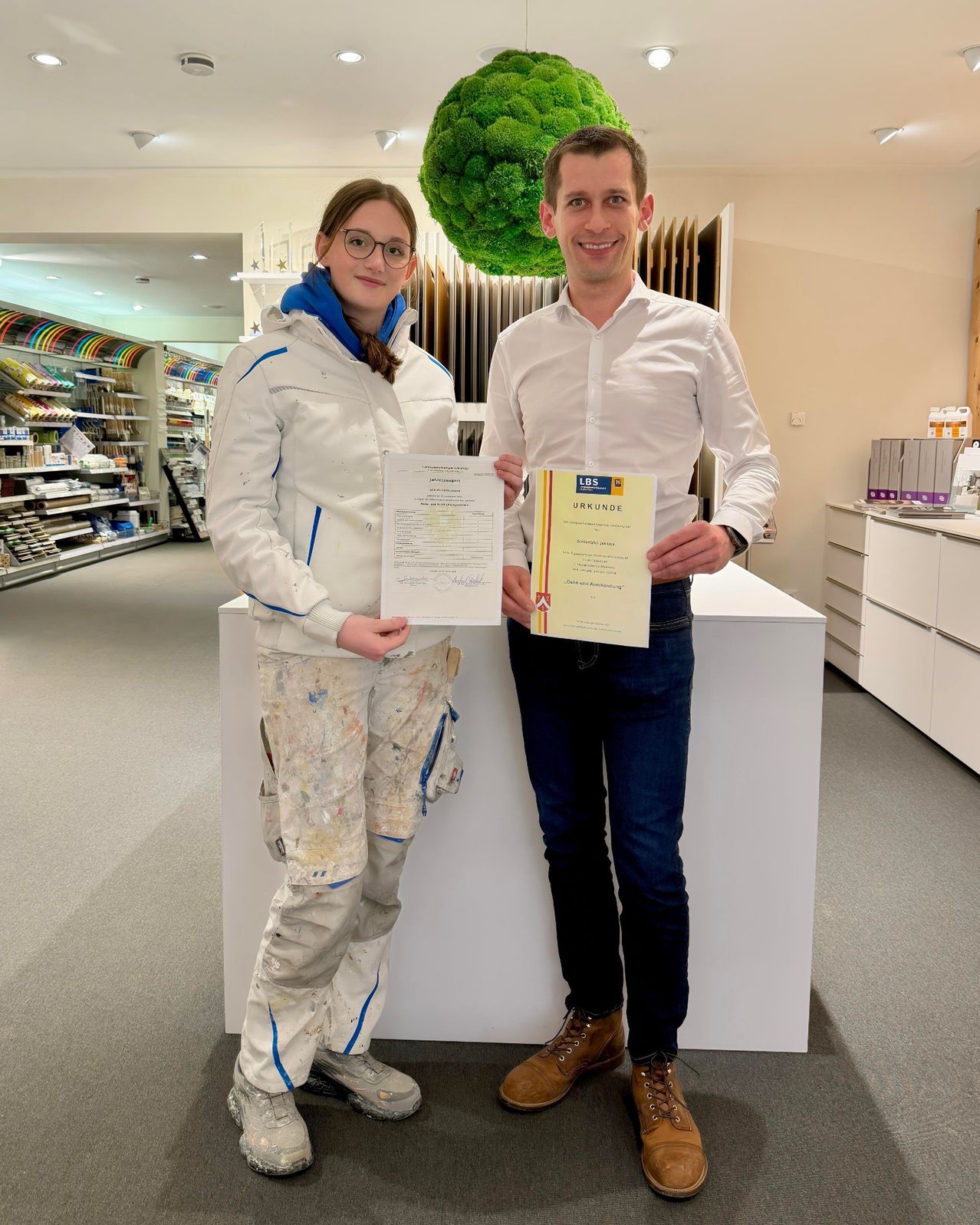 A woman and a man stand in a room, holding certificates, with a counter and shelves in the background.