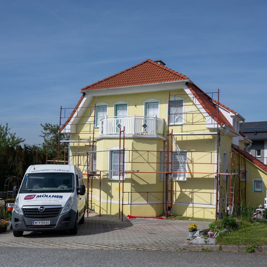 A yellow house with scaffolding around it, a white van parked in front, and a clear blue sky in the background.