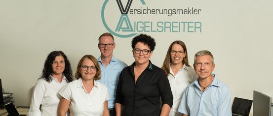 A group of six individuals, dressed in professional attire, are standing in front of a white wall with a logo. The woman in the center is wearing glasses and a black shirt.
