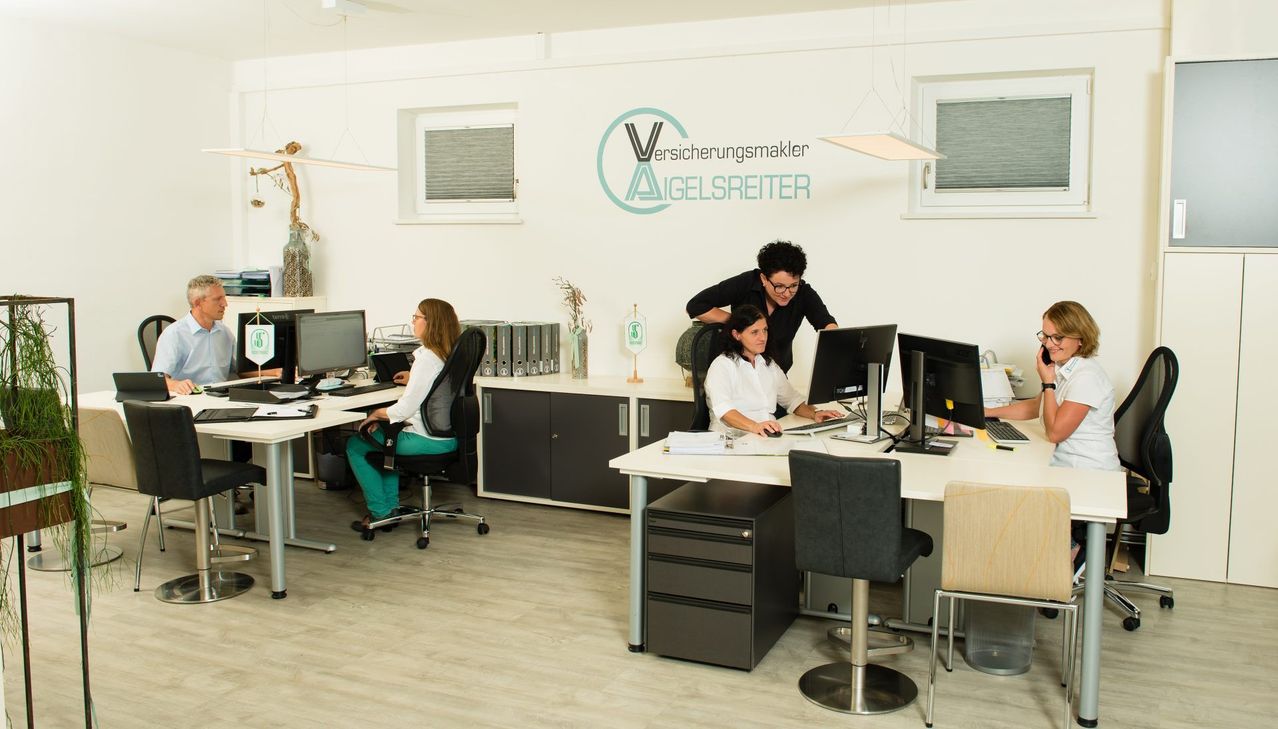 An office with three people working at computers. A man stands behind a woman at her desk. The office has a white wall with a logo and a window.