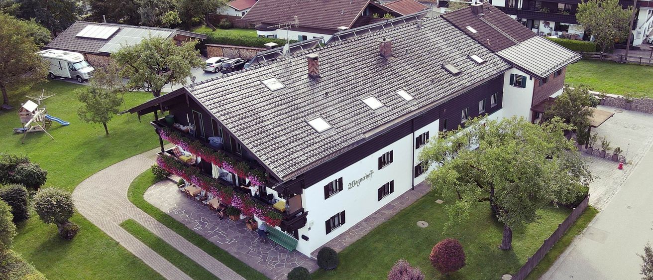 Eine Luftaufnahme eines großen weißen und braunen Gebäudes mit vielen Fenstern und einem braunen Dach. Bäume, Pflanzen und Gras umgeben es, vorn parken Autos.