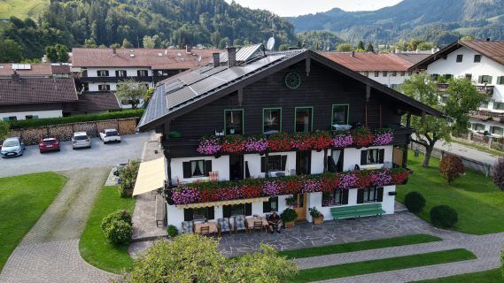 Ein Luftbild eines traditionellen Alpenhauses, geschmückt mit bunten Blumen und Solarpanelen. Das Haus hat eine große Einfahrt mit geparkten Autos und einem Garten.