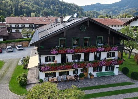 Ein Luftbild eines traditionellen Alpenhauses, geschmückt mit bunten Blumen und Solarpanelen. Das Haus hat eine große Einfahrt mit geparkten Autos und einem Garten.