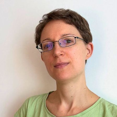 Eine Frau mit kurzen Haaren und Brille, die ein grünes T-Shirt trägt, steht vor einer weißen Wand und lächelt in die Kamera.