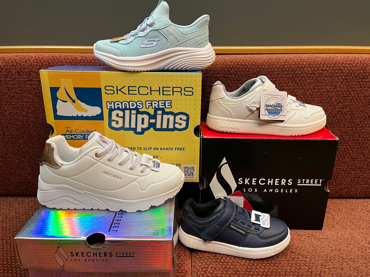 Vier Paar weiße, blaue und schwarze Skechers-Schuhe auf drei Kartons. Zwei Kartons haben 'Skechers' und 'Los Angeles' darauf geschrieben, einer ist gelb, der andere schwarz. Die oberen Schuhe sind türkis und weiß, die unteren marineblau und weiß.