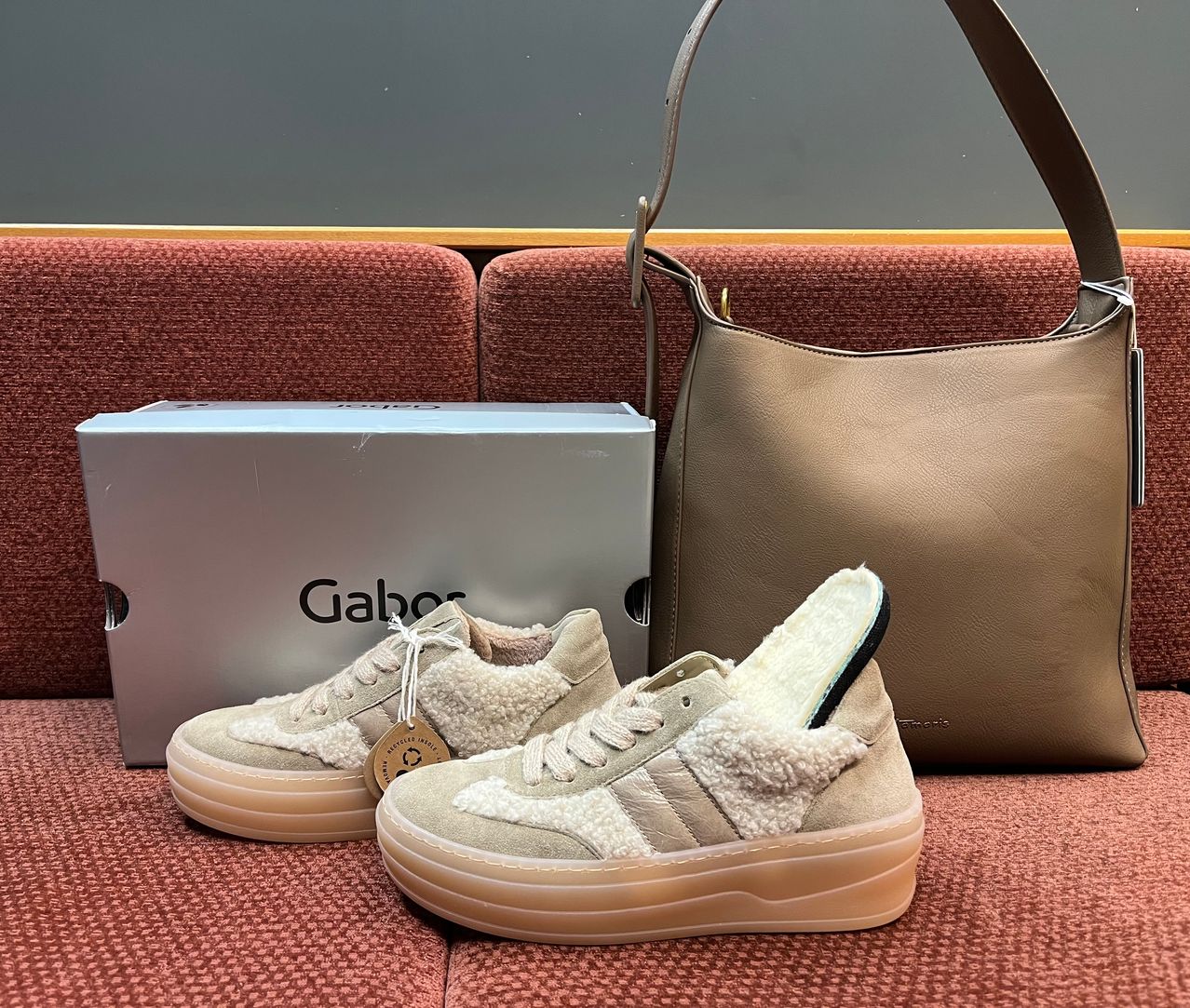 Ein Paar beige und weiße Sneakers mit Fellfutter und eine beige Handtasche liegen neben einer weißen Box mit dem Wort 'Gabor' darauf.