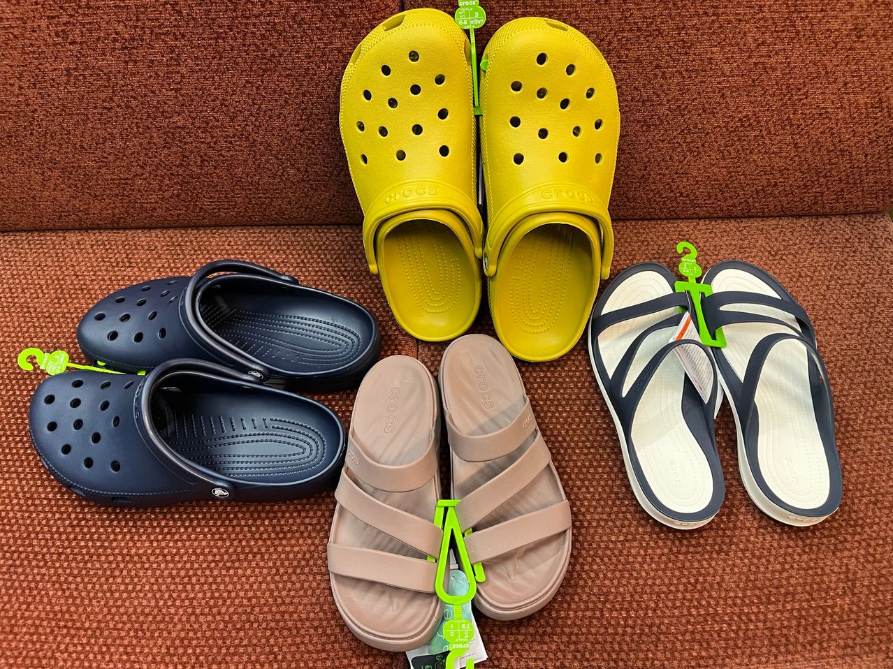 Eine Sammlung von Crocs-Sandalen und -Slides in verschiedenen Farben, einschließlich Gelb, Marineblau und Beige, wird auf einer braunen Oberfläche ausgestellt.