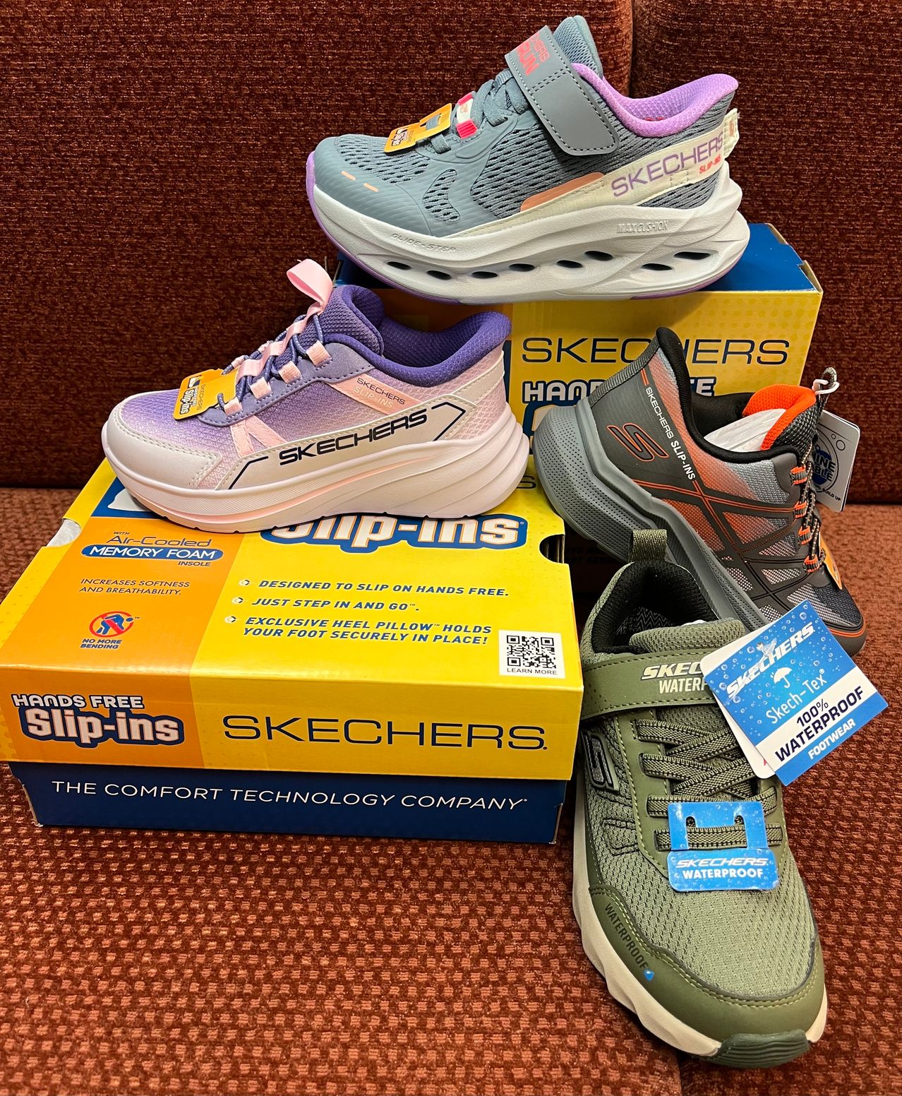 Vier Paare Skechers Slip-ins werden in Kartons ausgestellt. Ein Paar ist grau, ein anderes ist lila und weiß, ein weiteres ist grau und orange und das letzte ist grün.