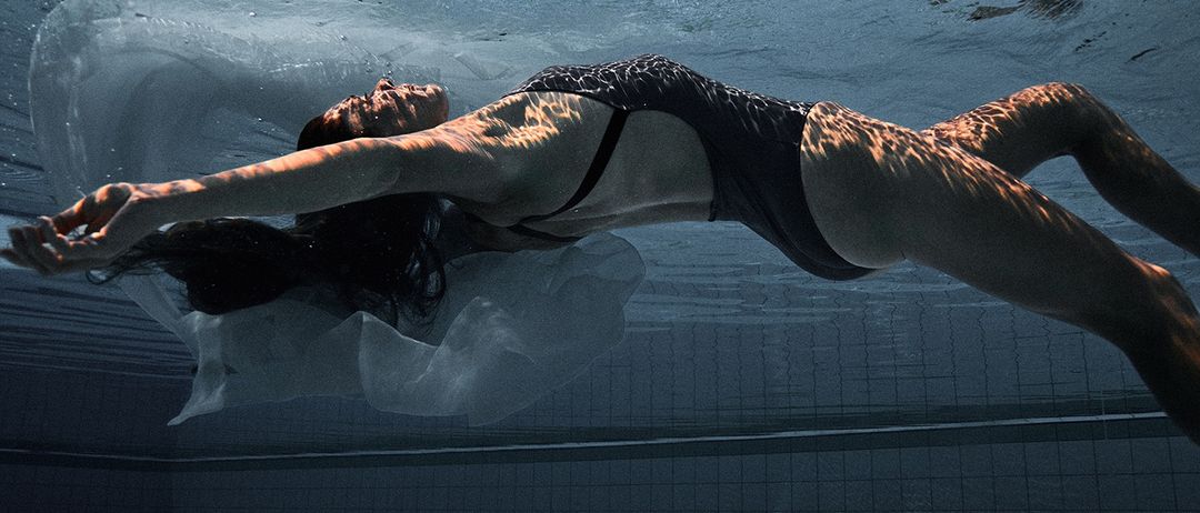 Eine Frau schwimmt anmutig unter Wasser in einem Pool, ihr Körper von weichem Licht beleuchtet, trägt einen schwarzen Badeanzug.