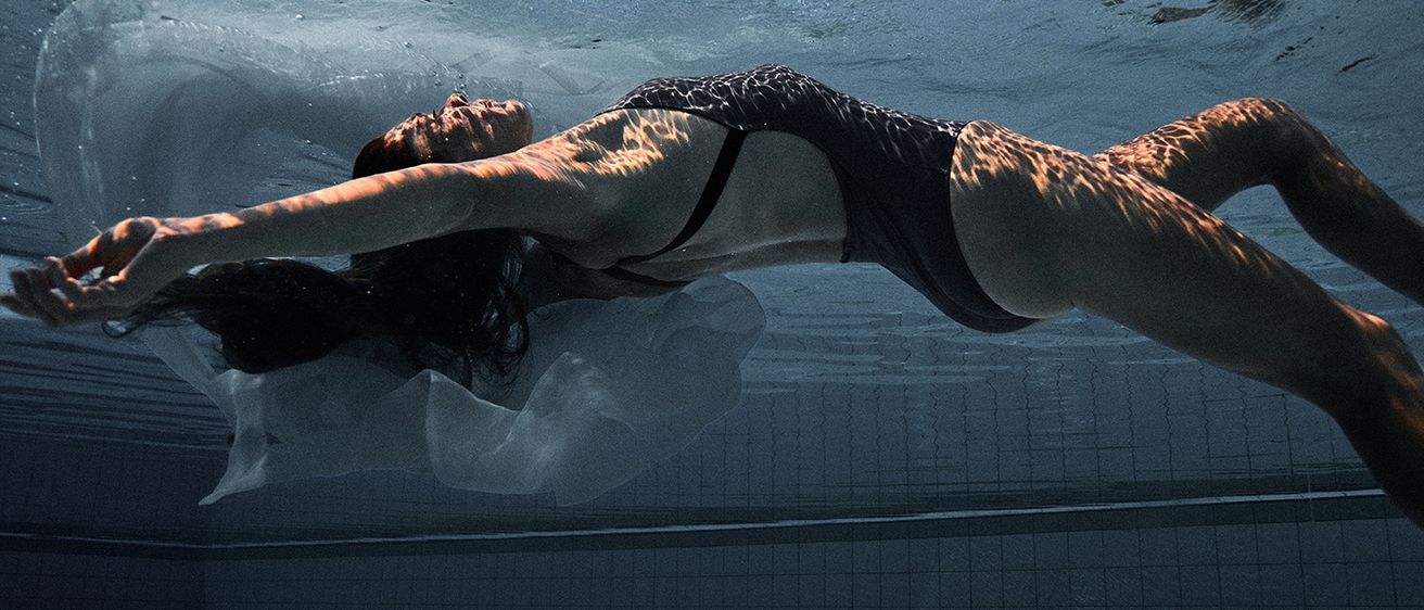 Eine Frau schwimmt anmutig unter Wasser in einem Pool, ihr Körper von weichem Licht beleuchtet, trägt einen schwarzen Badeanzug.