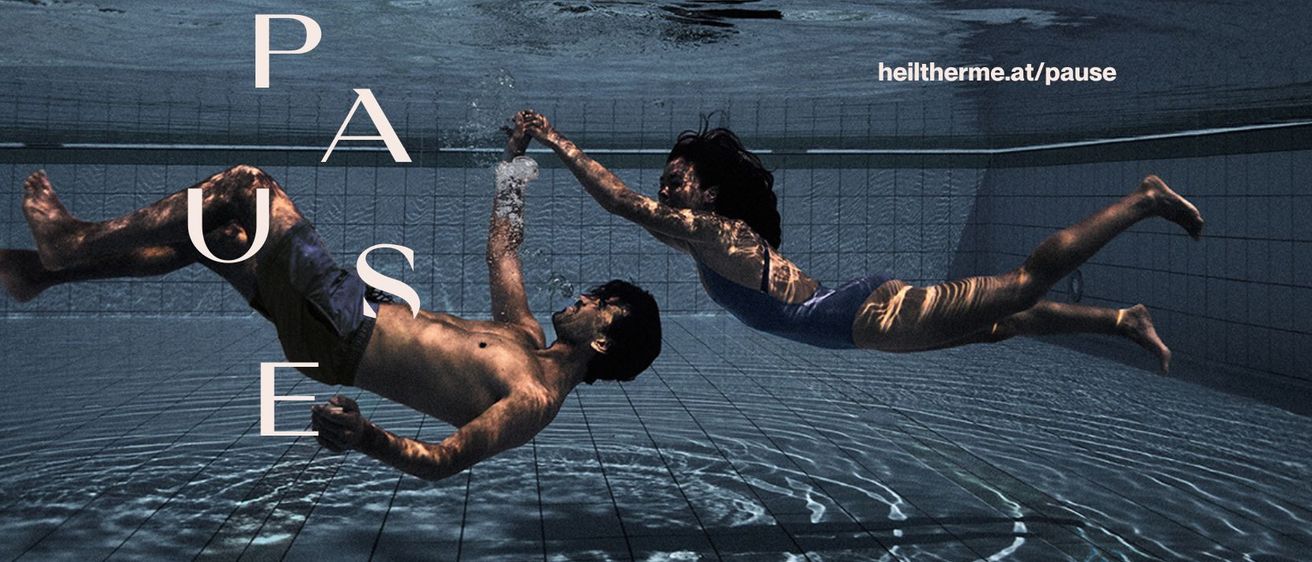 Zwei Personen schwimmen in einem Pool, eine auf dem Rücken und hält Hände, die andere auf dem Bauch, beide in Badekleidung.