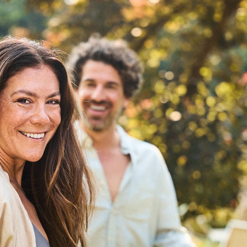 Ein Mann und eine Frau stehen nah beieinander und lächeln in die Kamera vor einem unscharfen Hintergrund aus Bäumen.