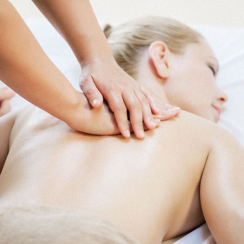 Eine Frau liegt auf dem Bauch auf einem Massagetisch. Eine Person massiert ihren Rücken mit der Hand.