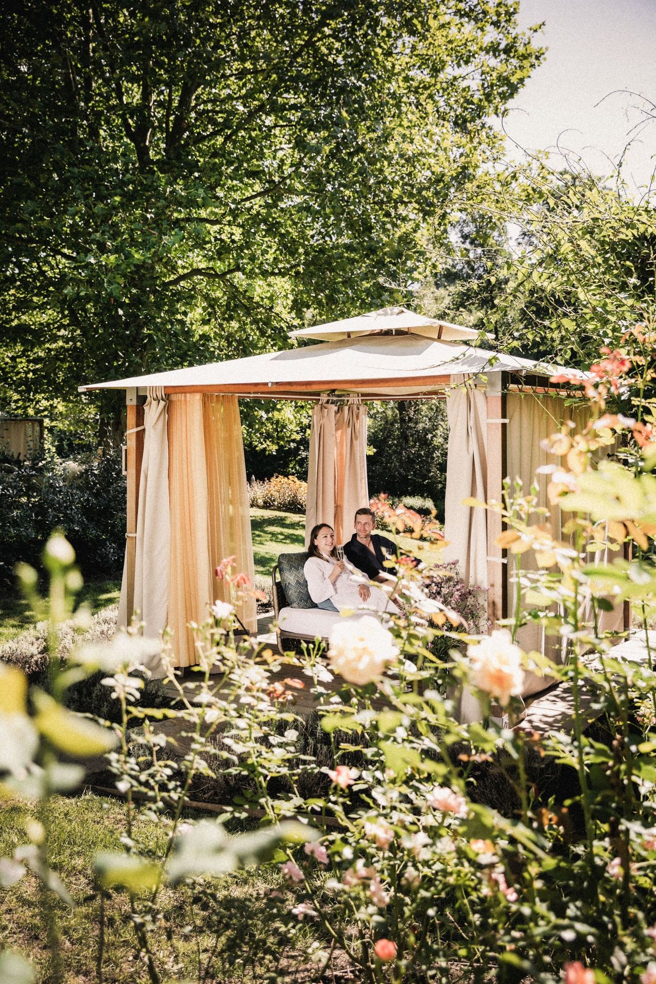 Ein Paar sitzt auf einem Liegestuhl unter einem Pavillon, umgeben von blühenden Blumen und Bäumen in einem Garten.