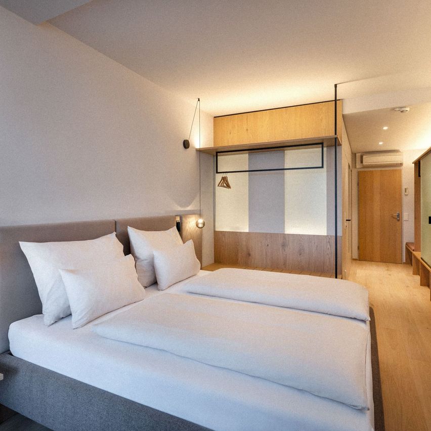 Ein modernes Schlafzimmer mit einem großen Bett, weißer Bettwäsche und mehreren weißen Kissen. Der Raum hat einen Holzboden und weiße Wände. Es gibt eine Holztür, die zu einem anderen Raum führt.