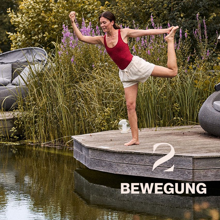 Eine Frau praktiziert Yoga auf einer Holzterrasse am Teich, umgeben von üppiger Grün- und Blumenlandschaft. Sie ist barfuß und trägt ein rotes Tanktop und weiße Shorts.